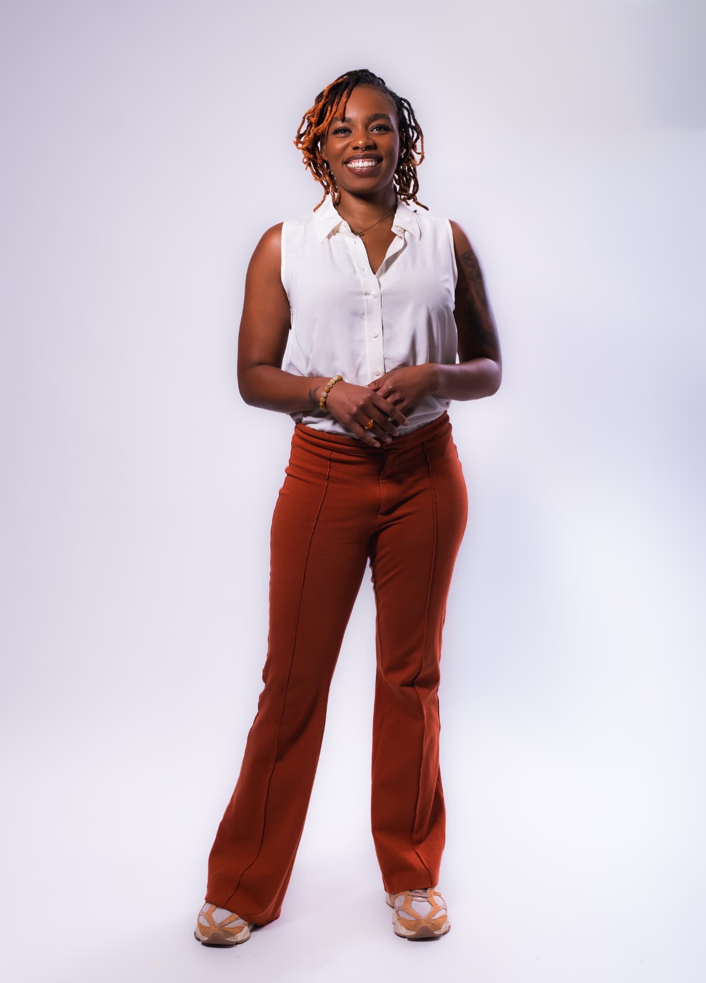 Smiling woman with orange locs in a white blouse and rust-colored pants on a white studio backdrop