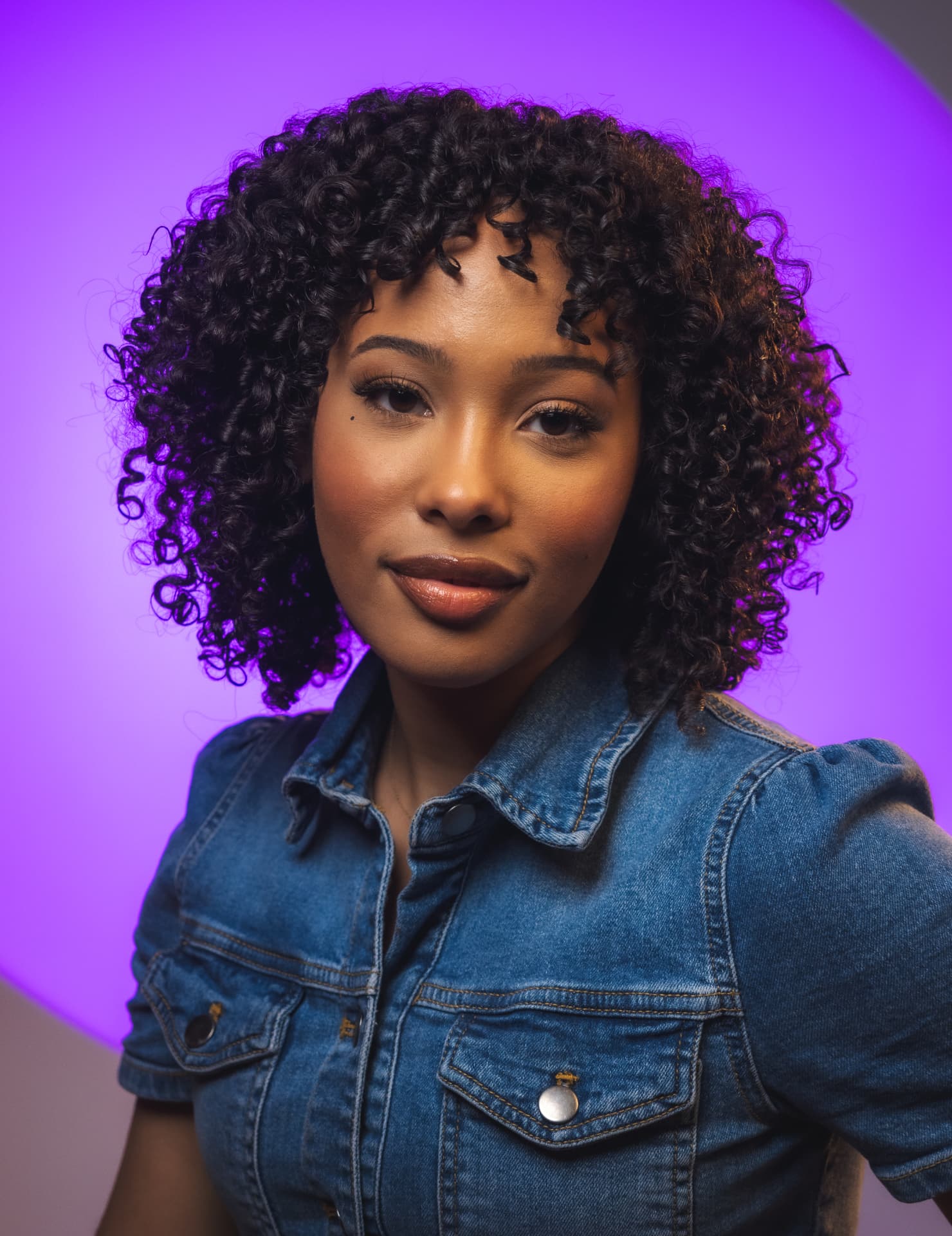 Smiling woman with curly hair in a denim jacket against a glowing purple circle backdrop