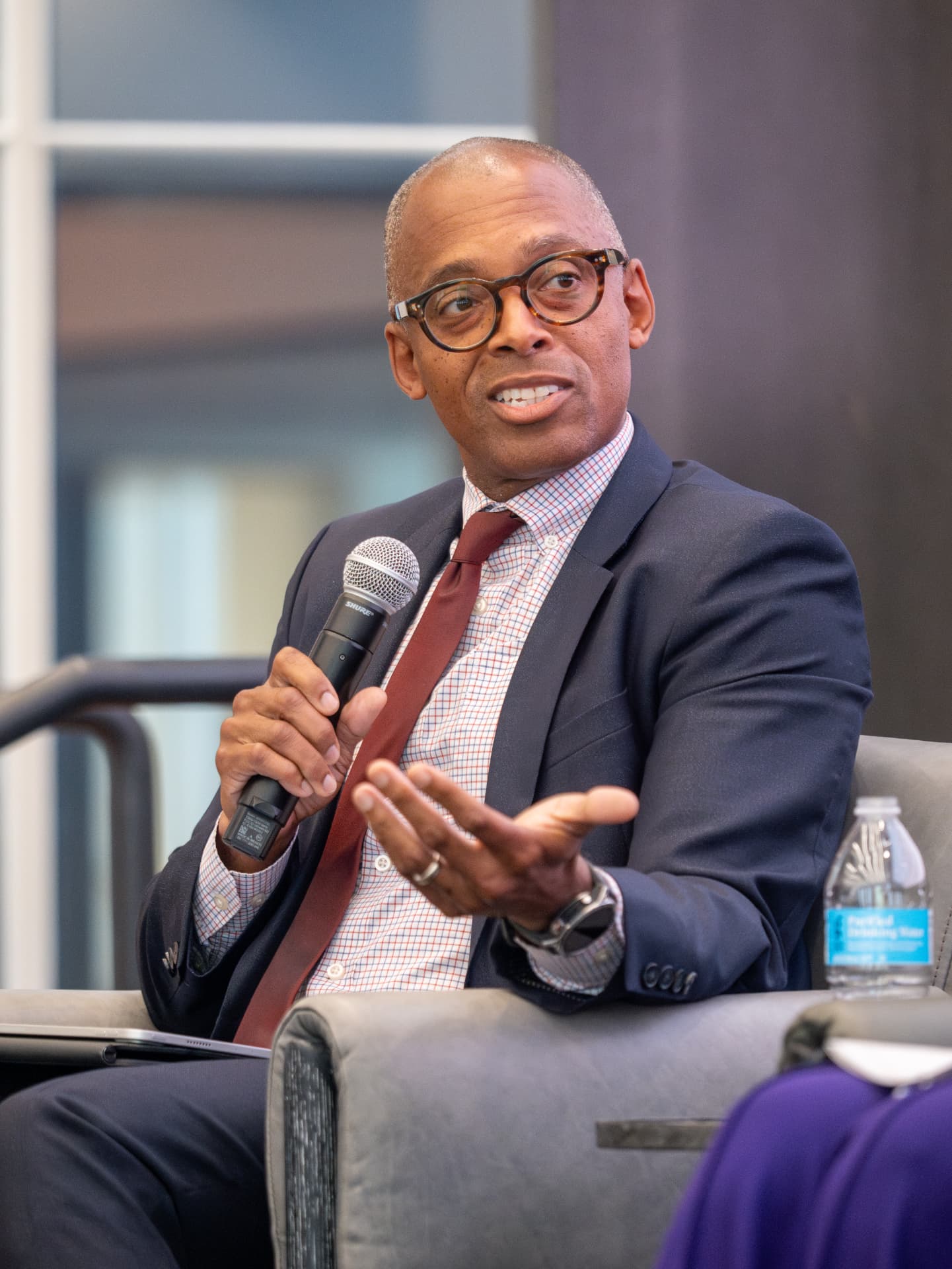 Man with glasses and a red tie speaking into a handheld microphone during a panel