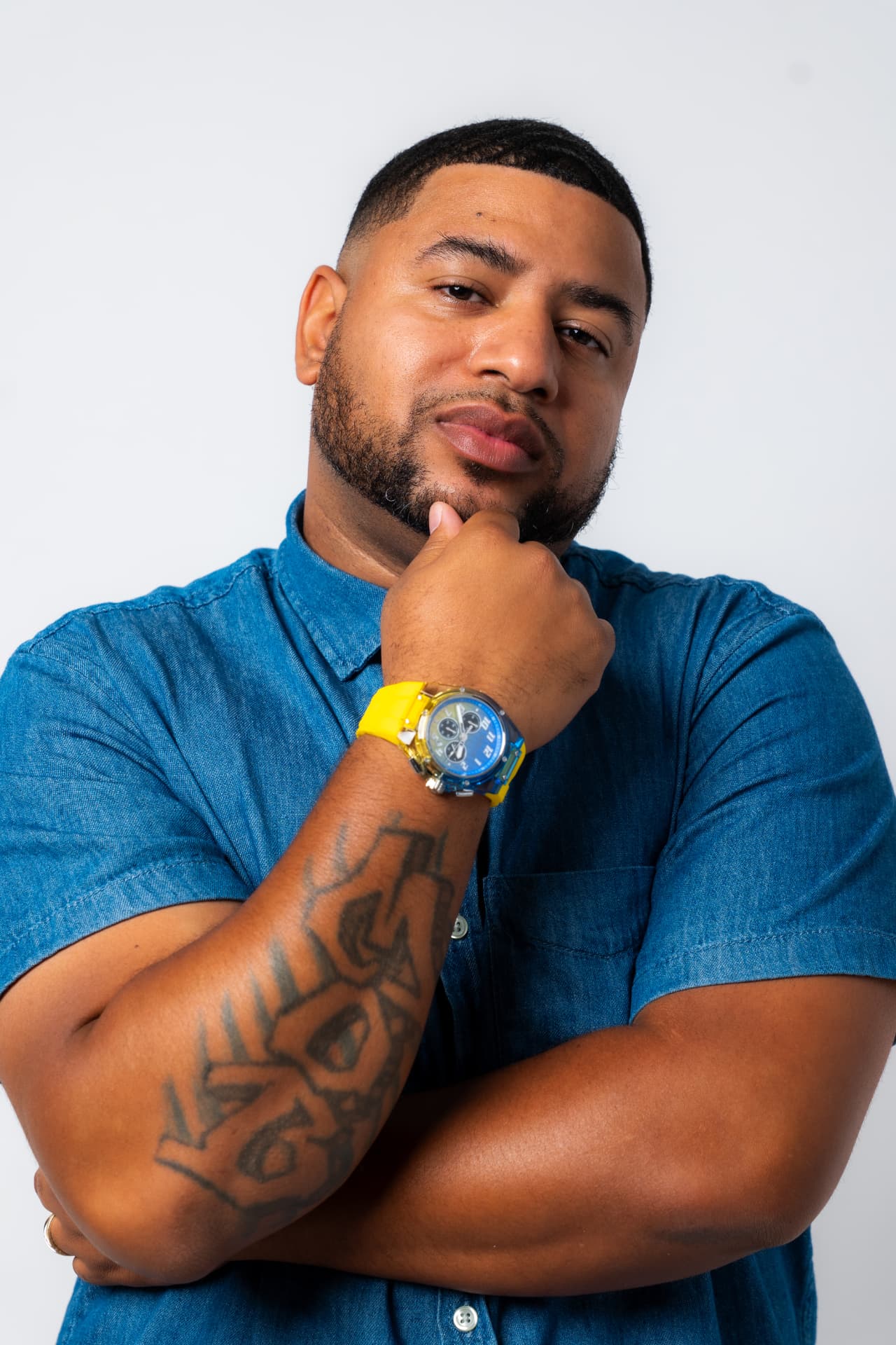 Man in a denim shirt with a yellow watch posing thoughtfully on a white studio backdrop