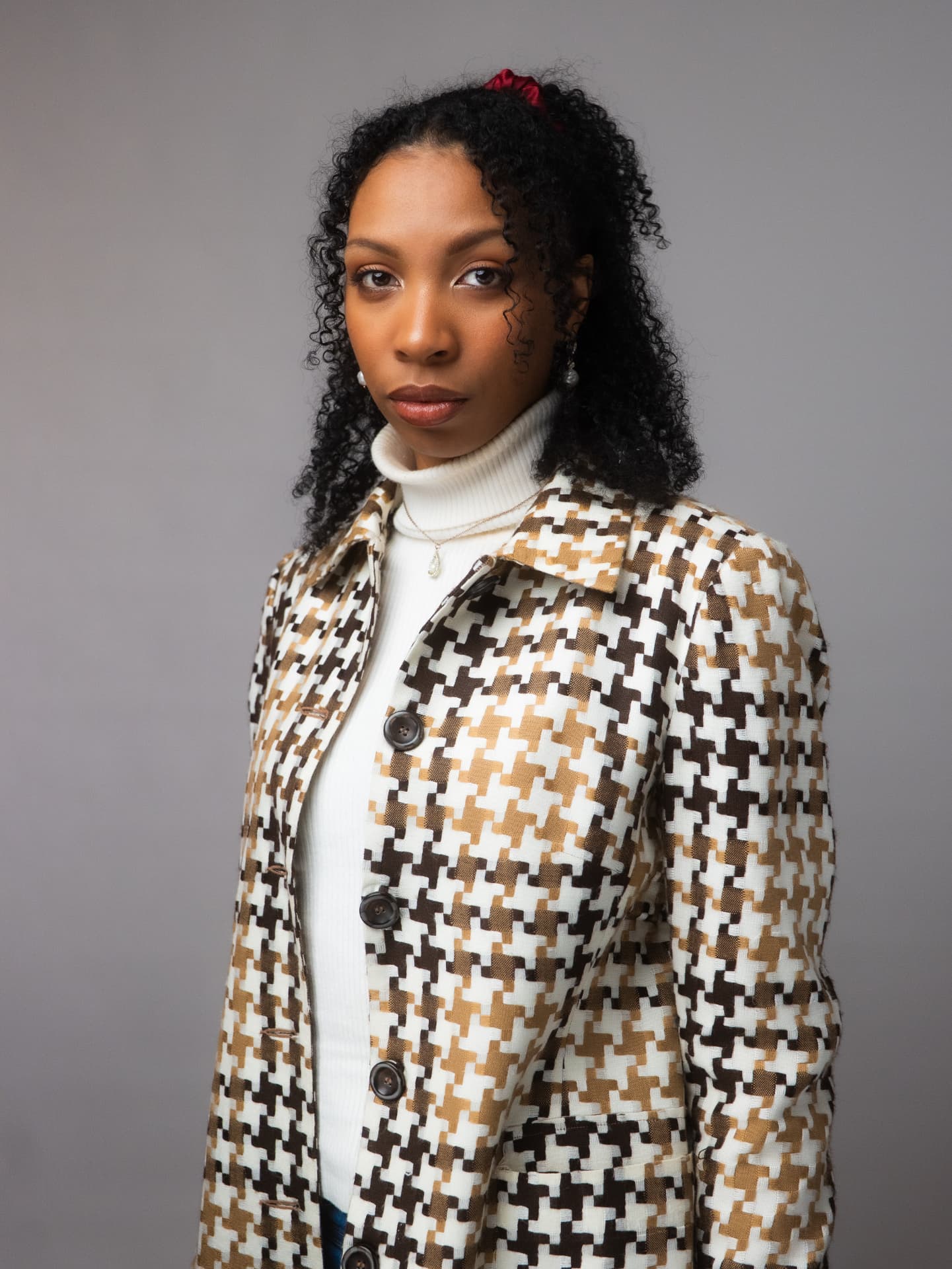 Woman in a houndstooth jacket over a white turtleneck posing against a gray backdrop