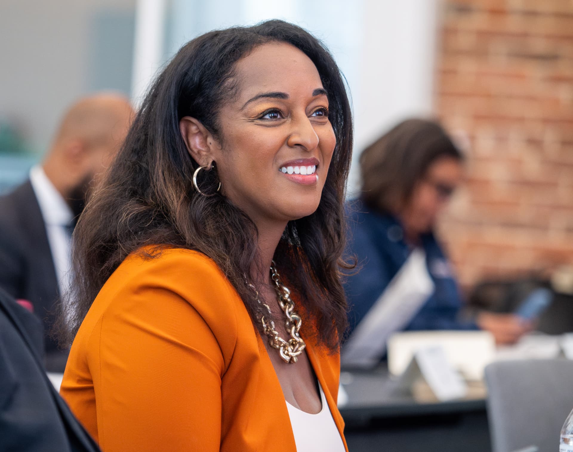 Woman in an orange blazer smiling at a conference table in front of a brick wall