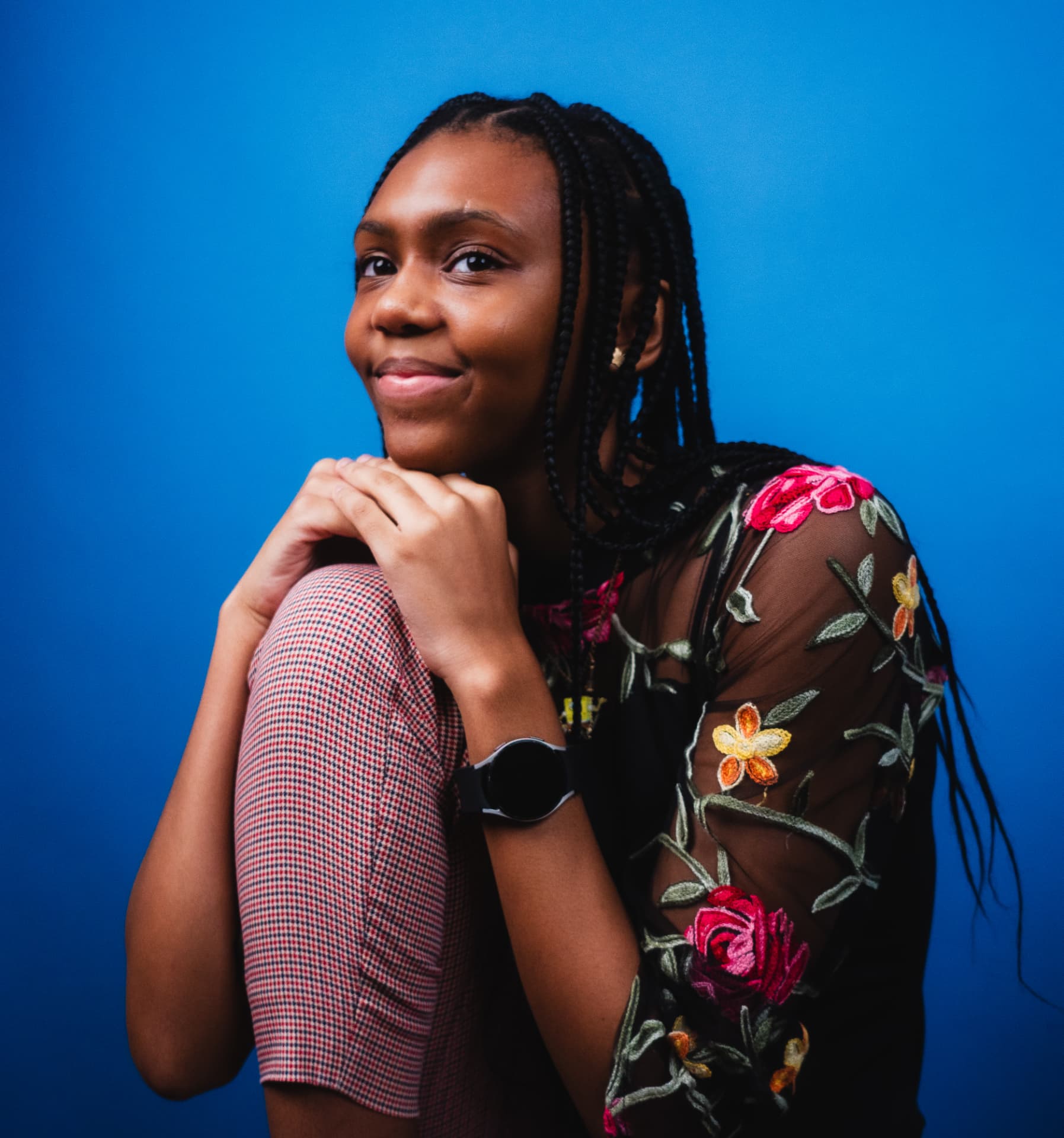 Woman with braids in a floral embroidered sheer top crouching against a blue background