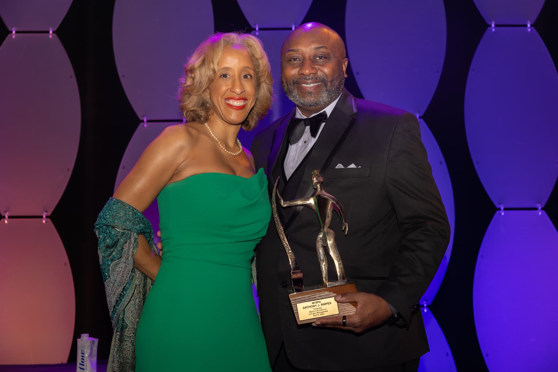Woman in a green gown and man in a tuxedo holding an award, posing together at a gala