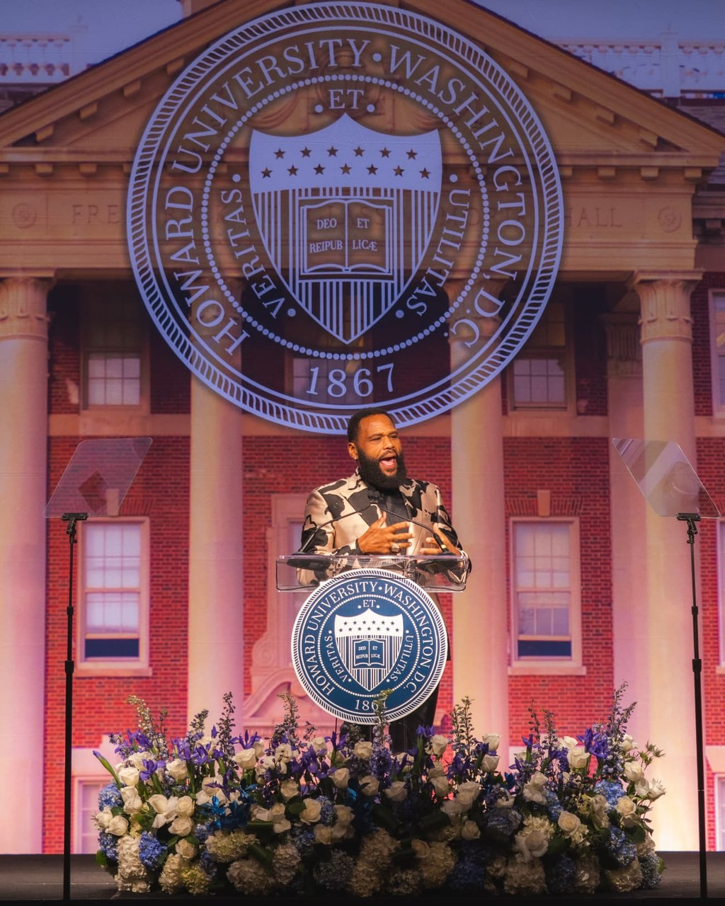 Speaker at a glass podium on stage in front of a large Howard University seal backdrop