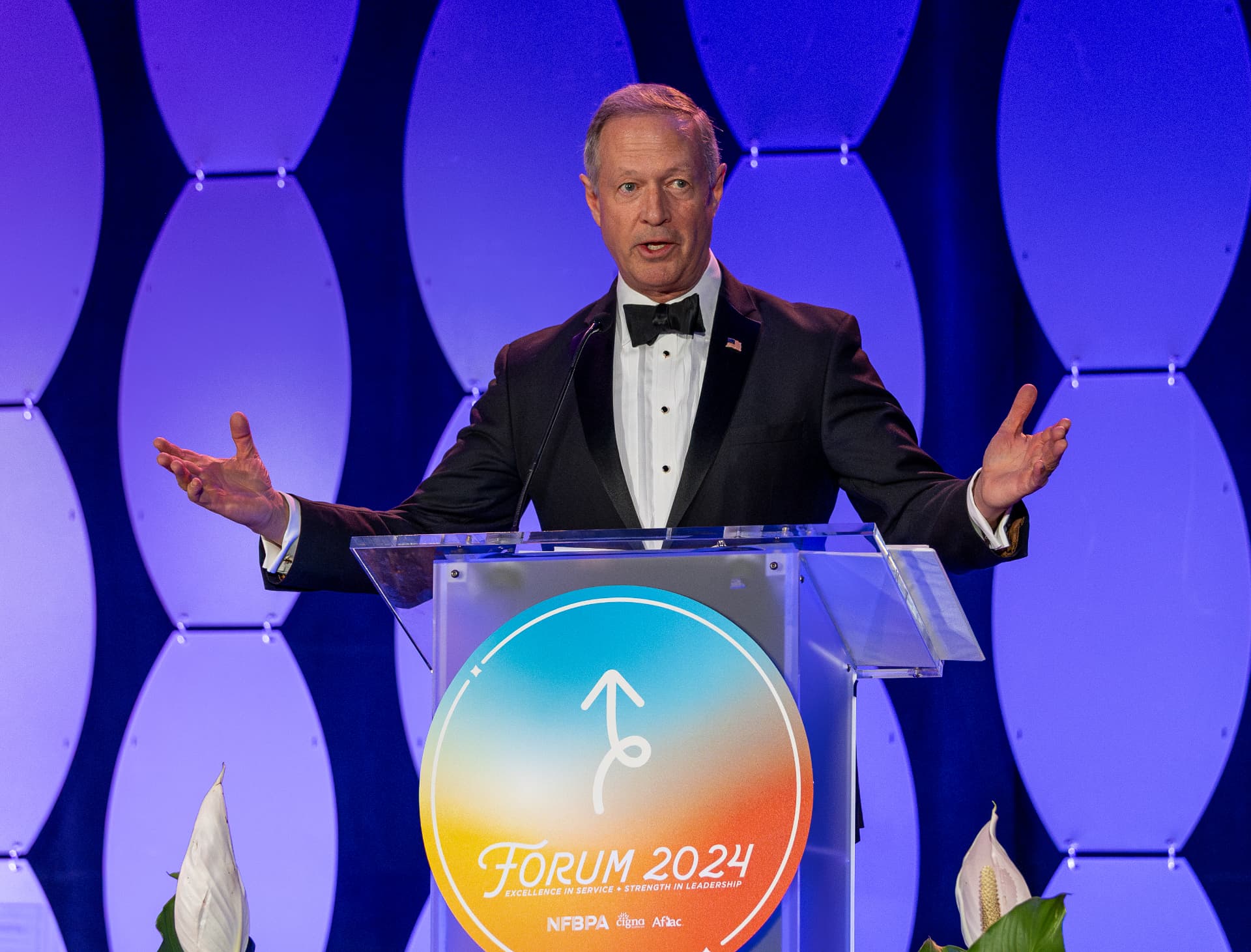 Man in a tuxedo speaking at the clear podium in front of the Forum 2024 medallion