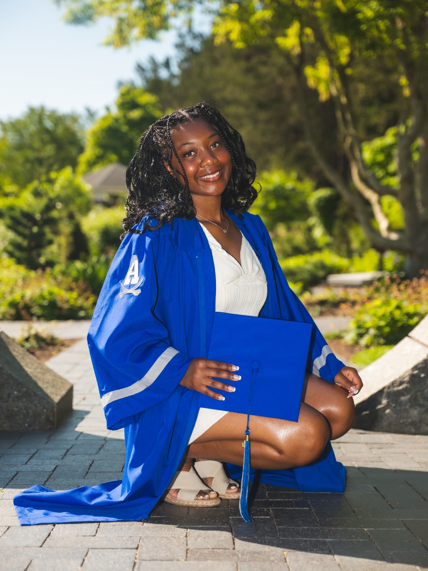 Graduate in a blue cap and gown kneeling in a sunlit garden holding her diploma cap