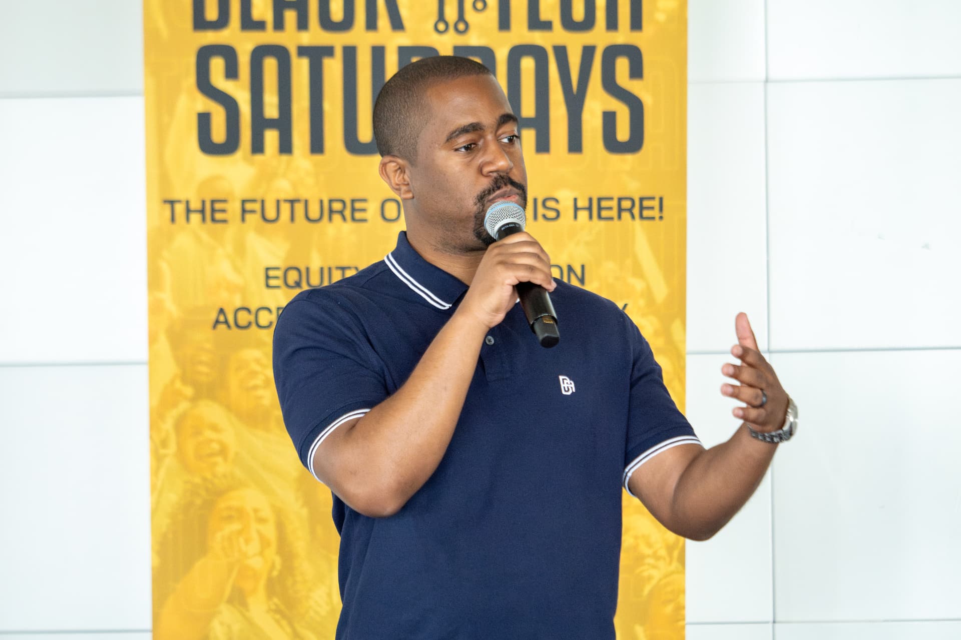 Man in a navy polo speaking into a microphone in front of the Black Tech Saturdays banner