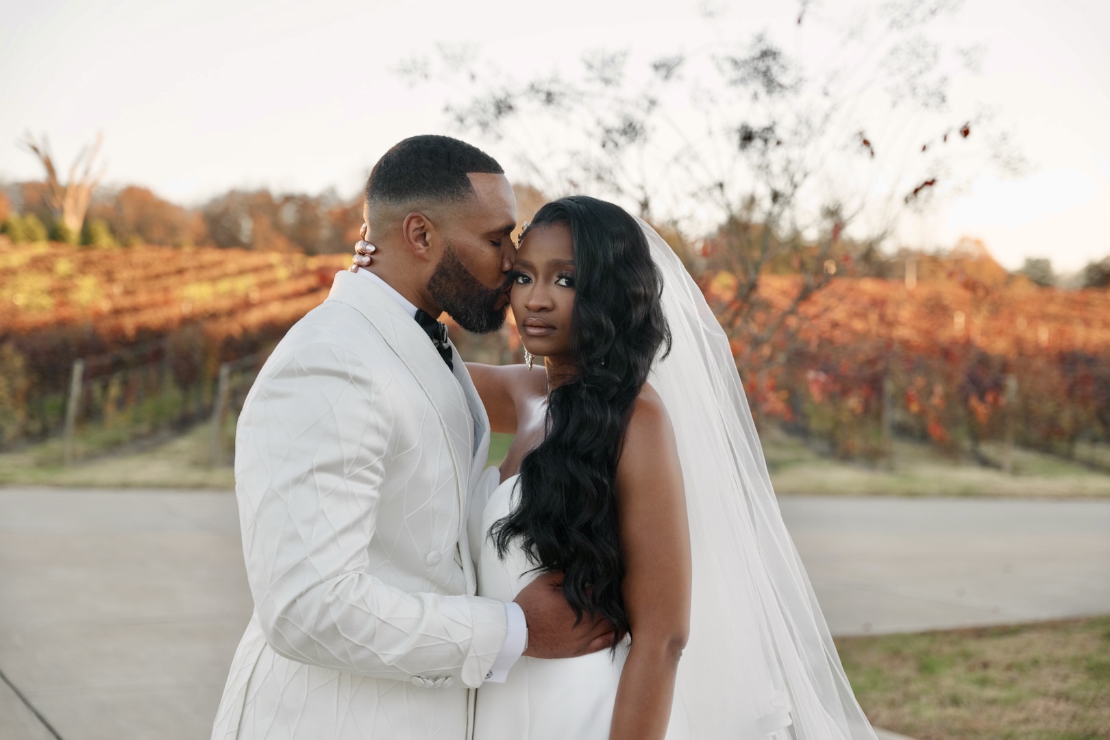 Groom kissing the bride's temple as she gazes at the camera in a vineyard at golden hour