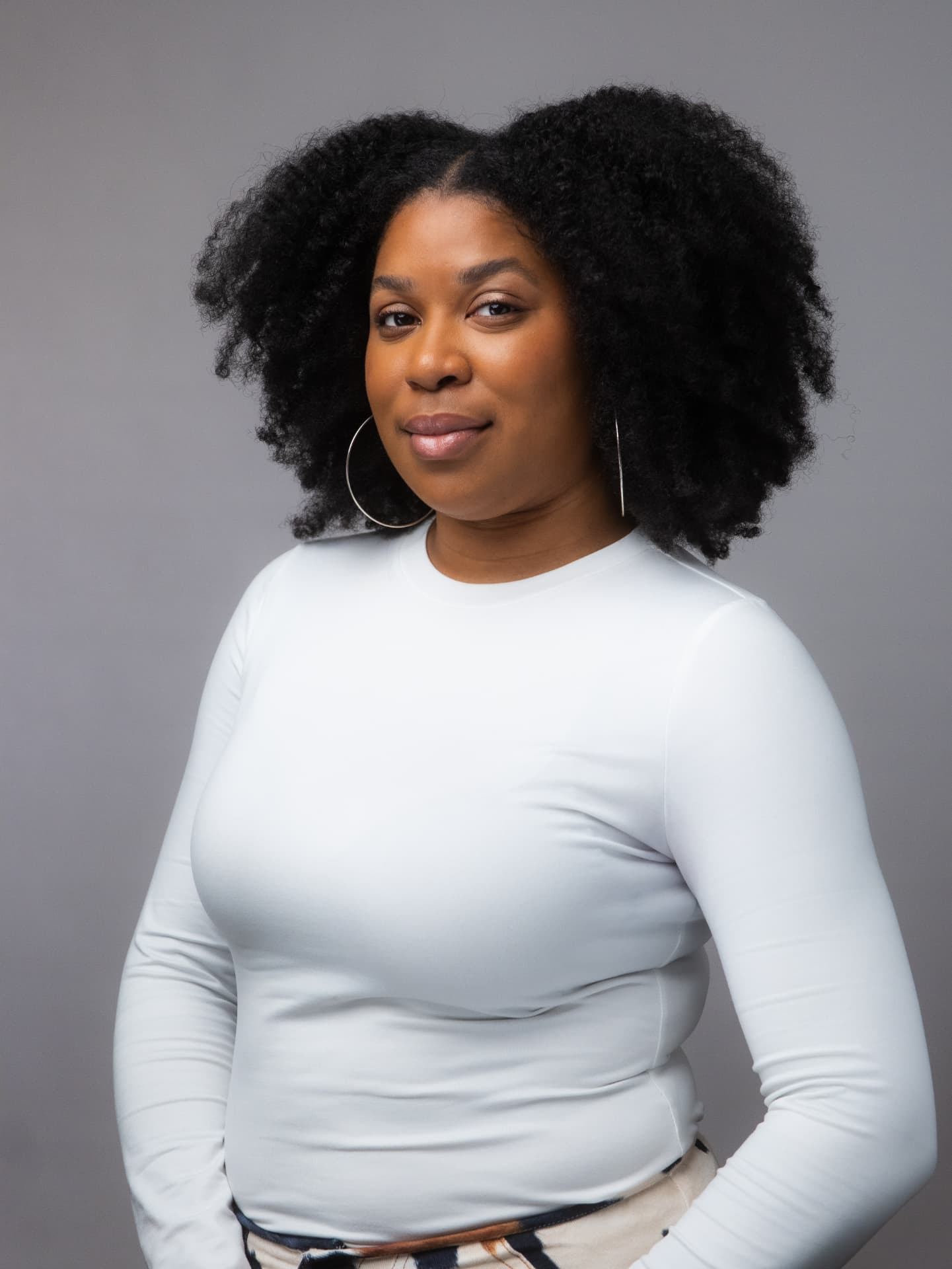 Woman with voluminous curls and silver hoop earrings in a white top against a gray backdrop