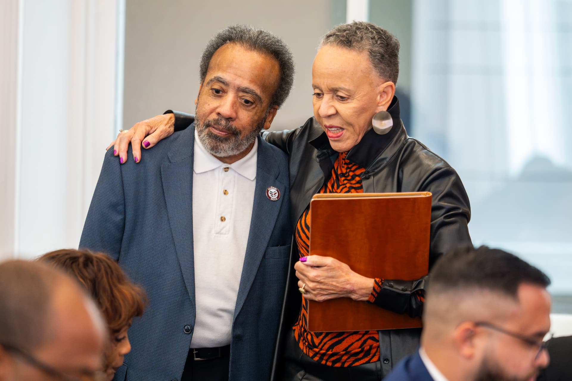 Woman in a leather jacket with her arm around a man in a blue blazer holding a wooden folder