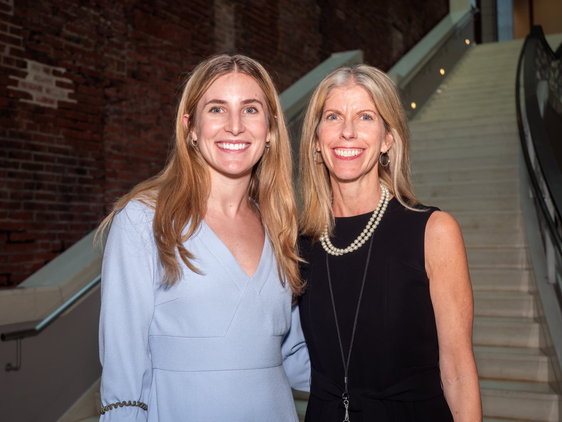 Two women in dresses posing near a spiral staircase at an evening event