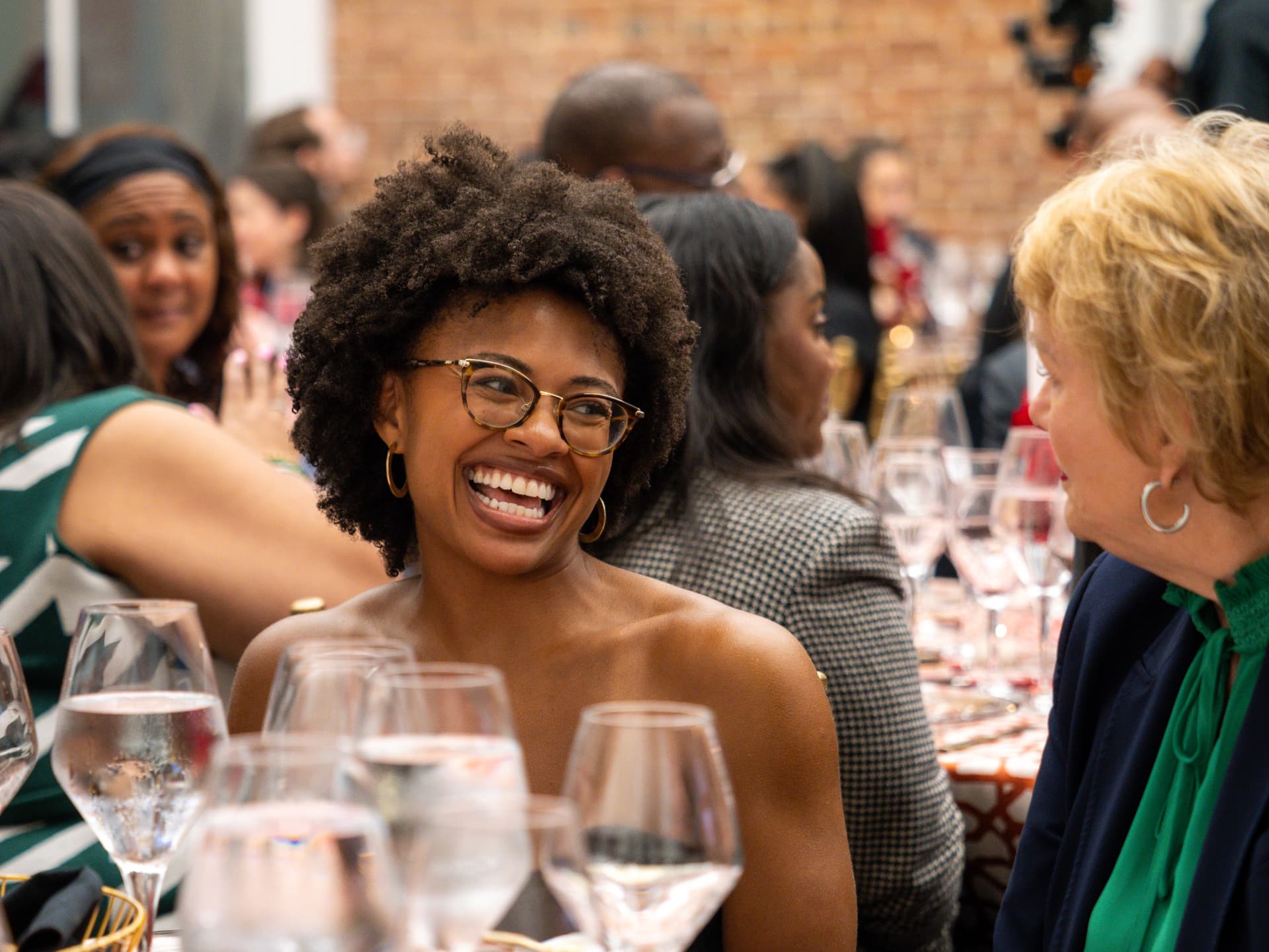 Woman with an afro and glasses laughing at a dinner table among wine glasses