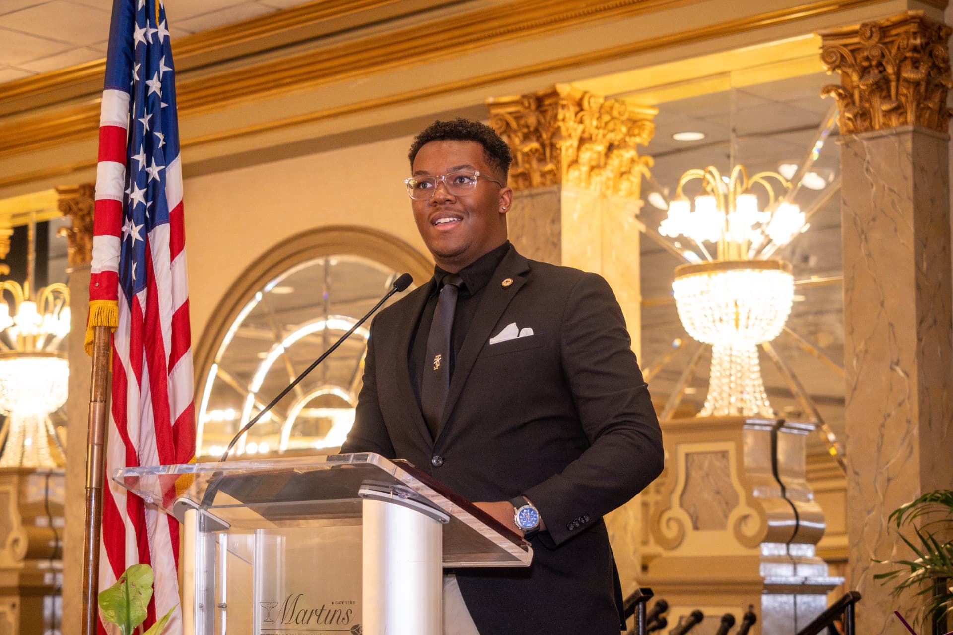 Young speaker at a podium beside an American flag in a gilded ballroom