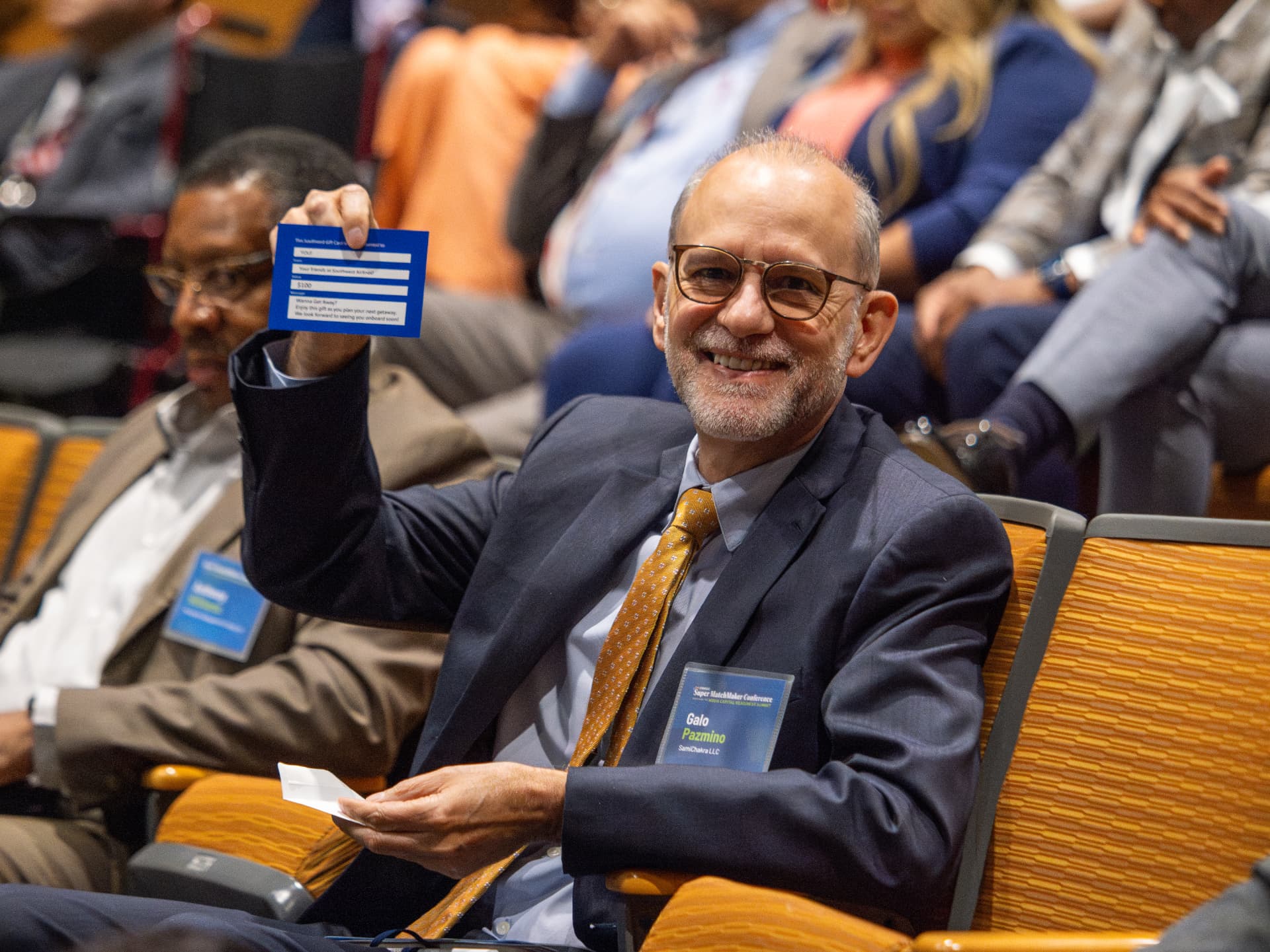 Man in glasses smiling and holding up a gift card while seated in an auditorium