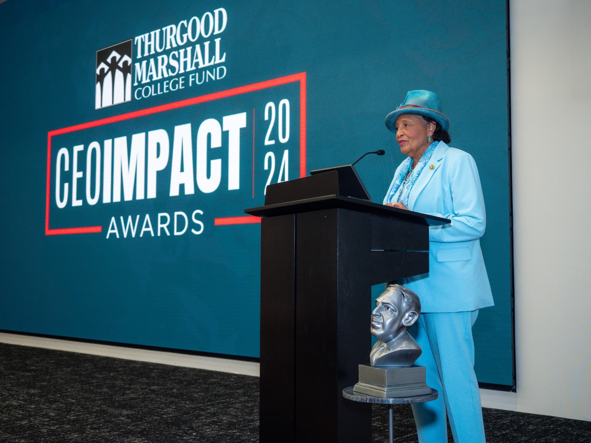 Woman in a light blue suit and hat speaking at the podium at the CEO IMPACT Awards