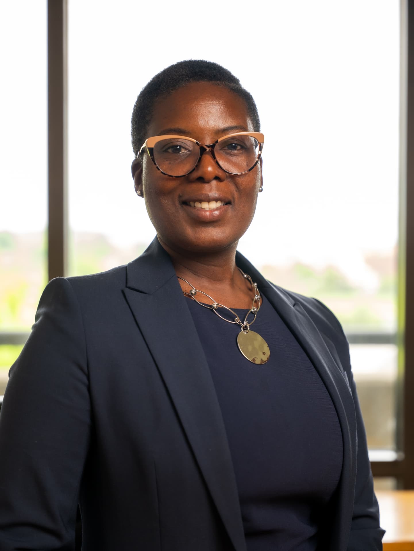Professional headshot of a woman in a navy suit and glasses beside an office window