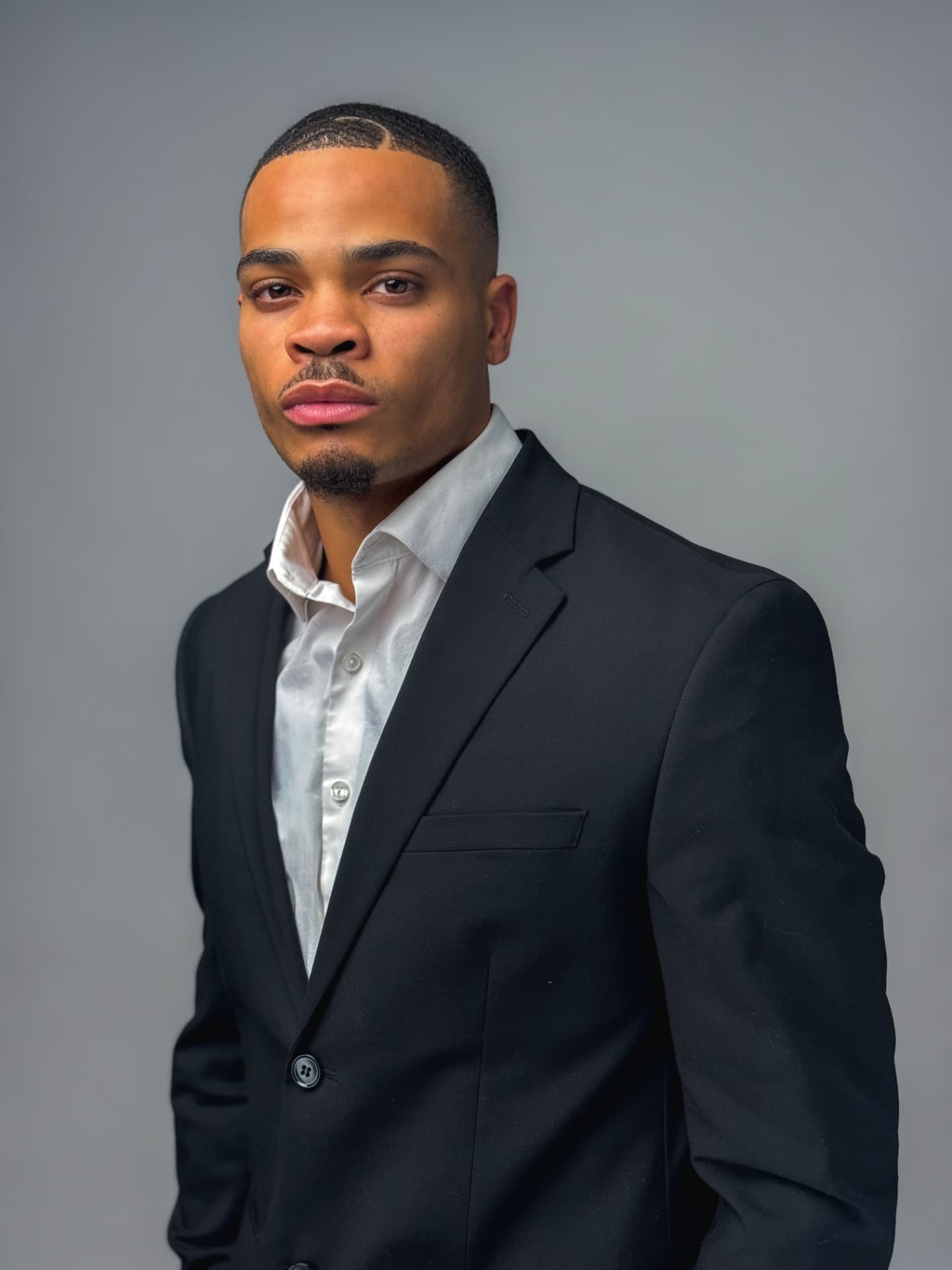 Man in a black suit and open white shirt posing against a gray studio backdrop
