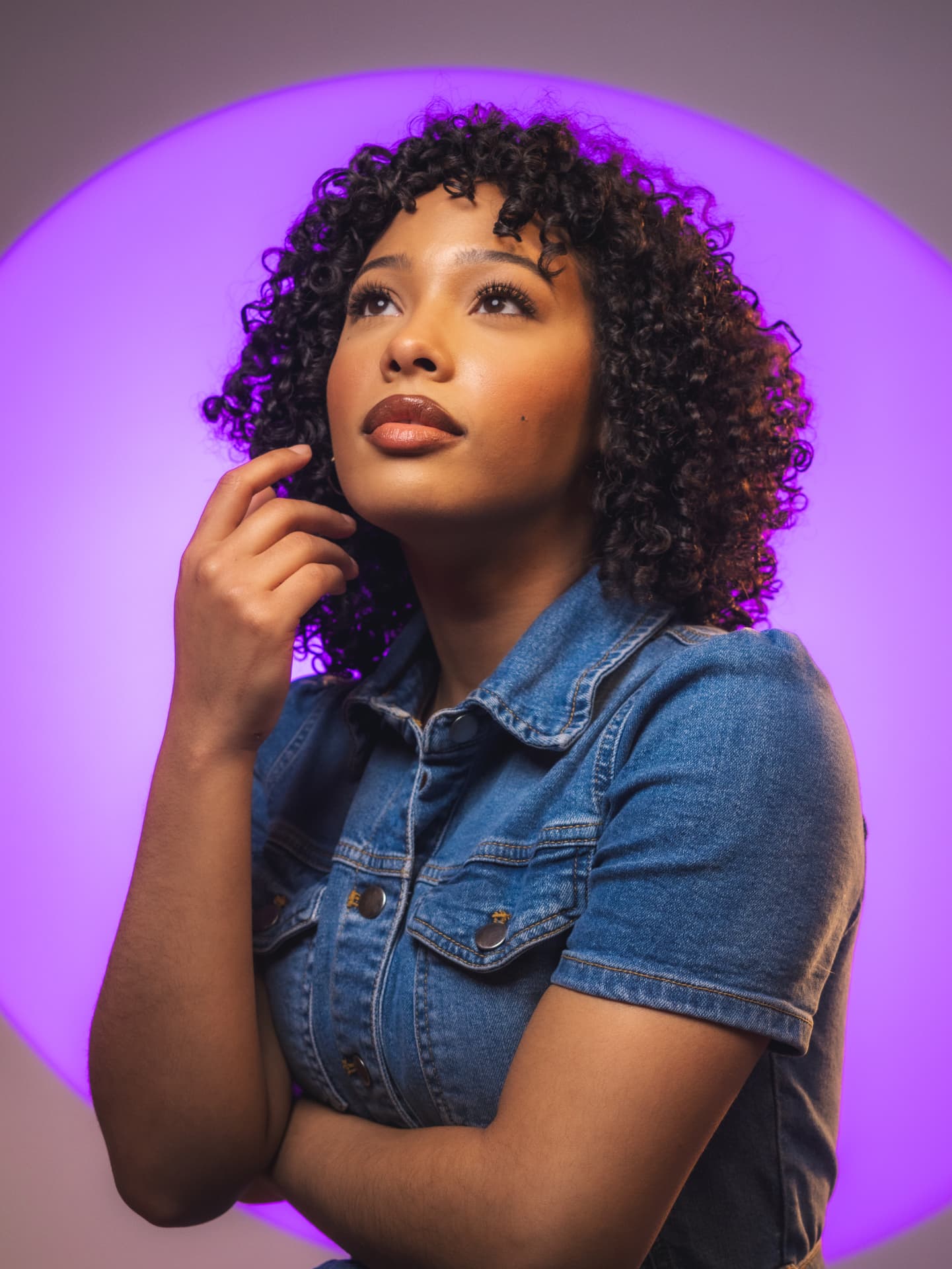 Woman with curly hair in a denim jacket gazing upward with a raised hand against a purple glow