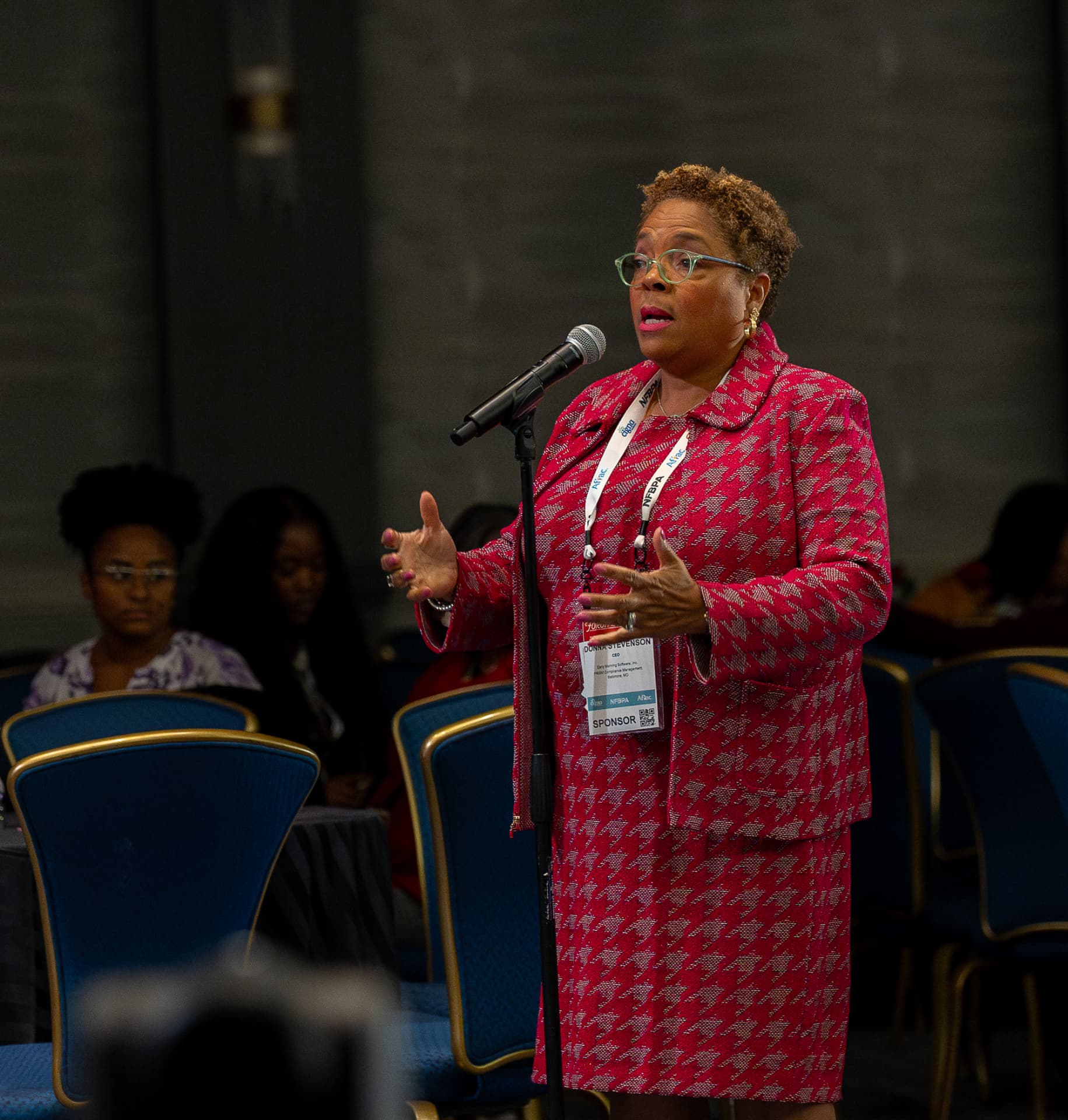 Woman in a red houndstooth suit addressing attendees at a conference session