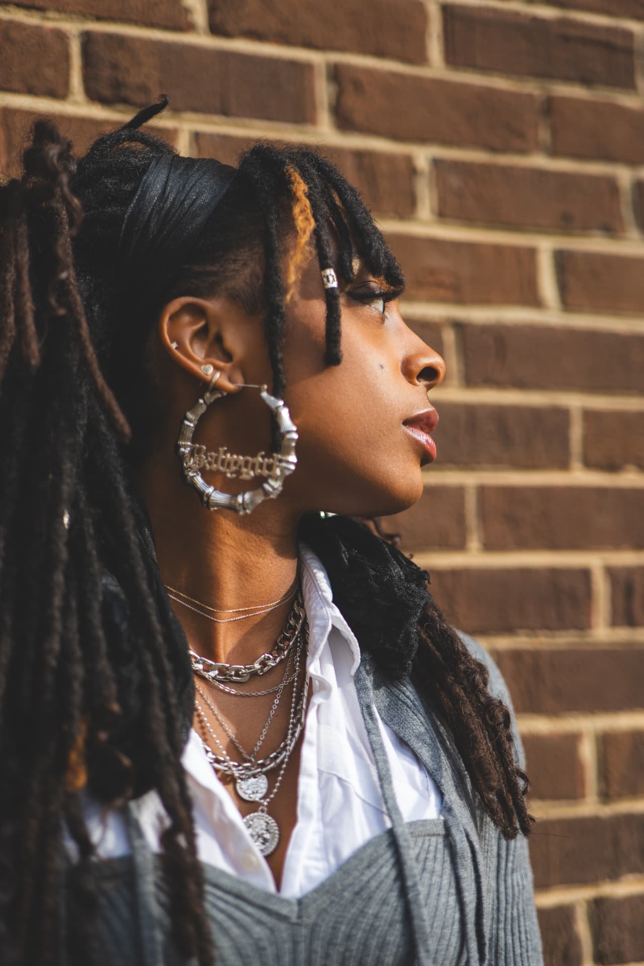 Profile of a woman with locs and bamboo hoop earrings wearing layered silver chains against a brick wall