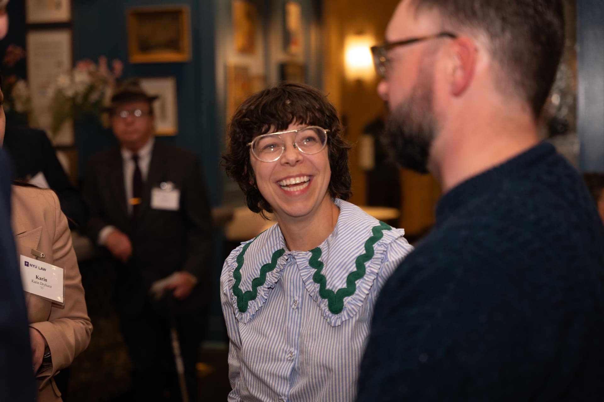 Woman with round glasses and a rickrack-trimmed collar laughing with a guest at a reception
